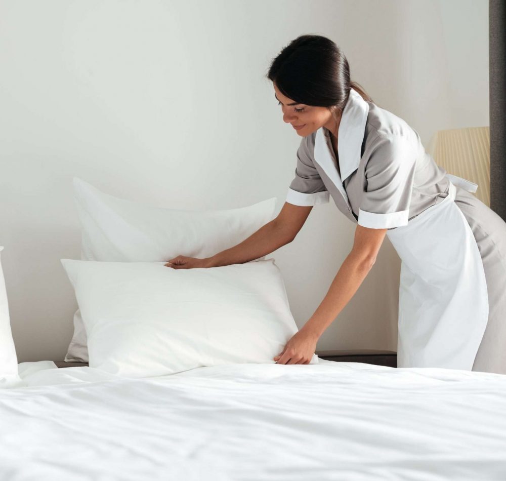 Young hotel maid setting up white pillow on bed sheet in hotel room
